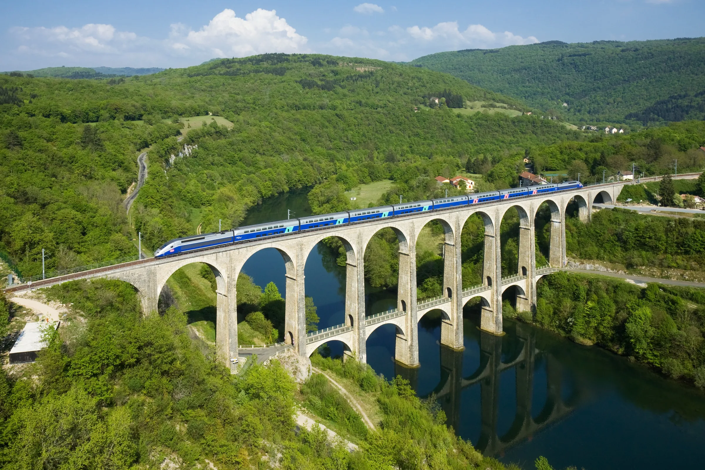 A blue SNCF TGV high-speed train crossing the Cize–Bolozon stone viaduct above a river